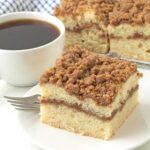 A slice of coffee cake on a plate, a cup of coffee sits to the left and more cake sits in the background.