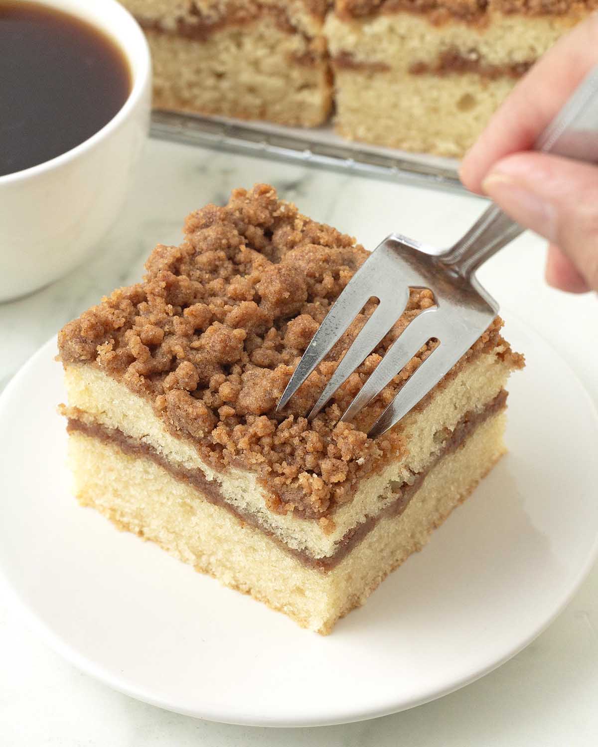 A hand holding a fork that is digging into a slice of coffee cake.