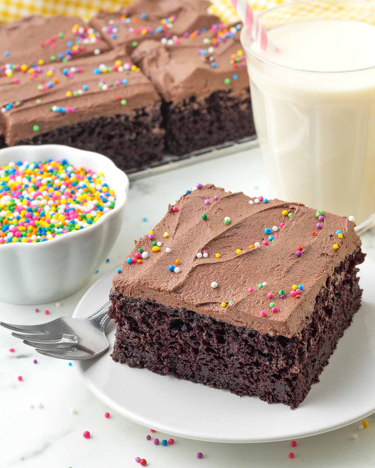A square of vegan chocolate sheet cake on a plate, the cake is frosted and topped with round rainbow sprinkles.