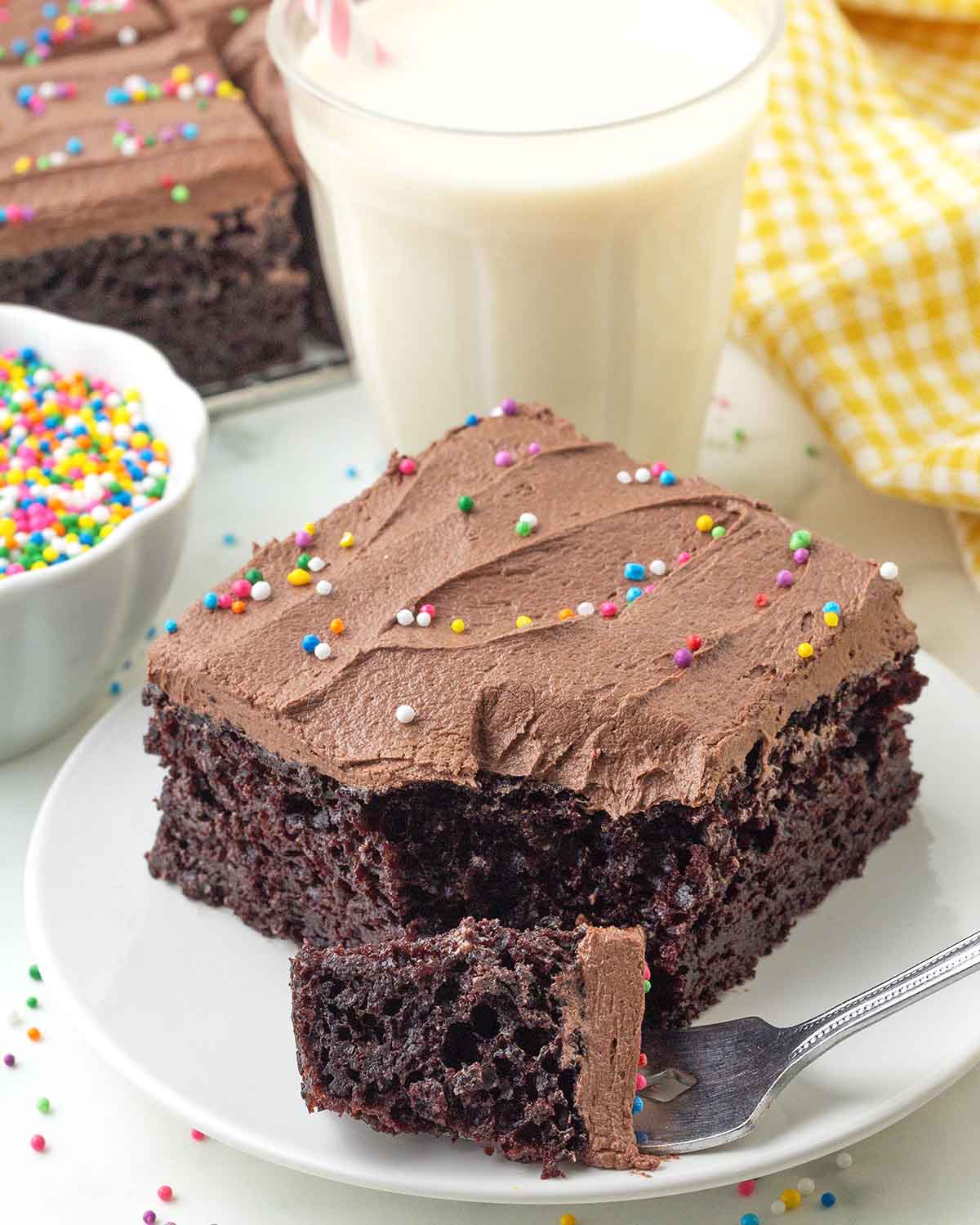 A piece of vegan chocolate sheet cake on a white plate, a fork with a piece of the cake on it sits in front of the cake.