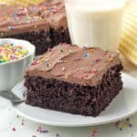 A slice of vegan chocolate sheet cake on a plate, a glass of milk sits behind the plate, a bowl of sprinkles to the left and more cake in the background.