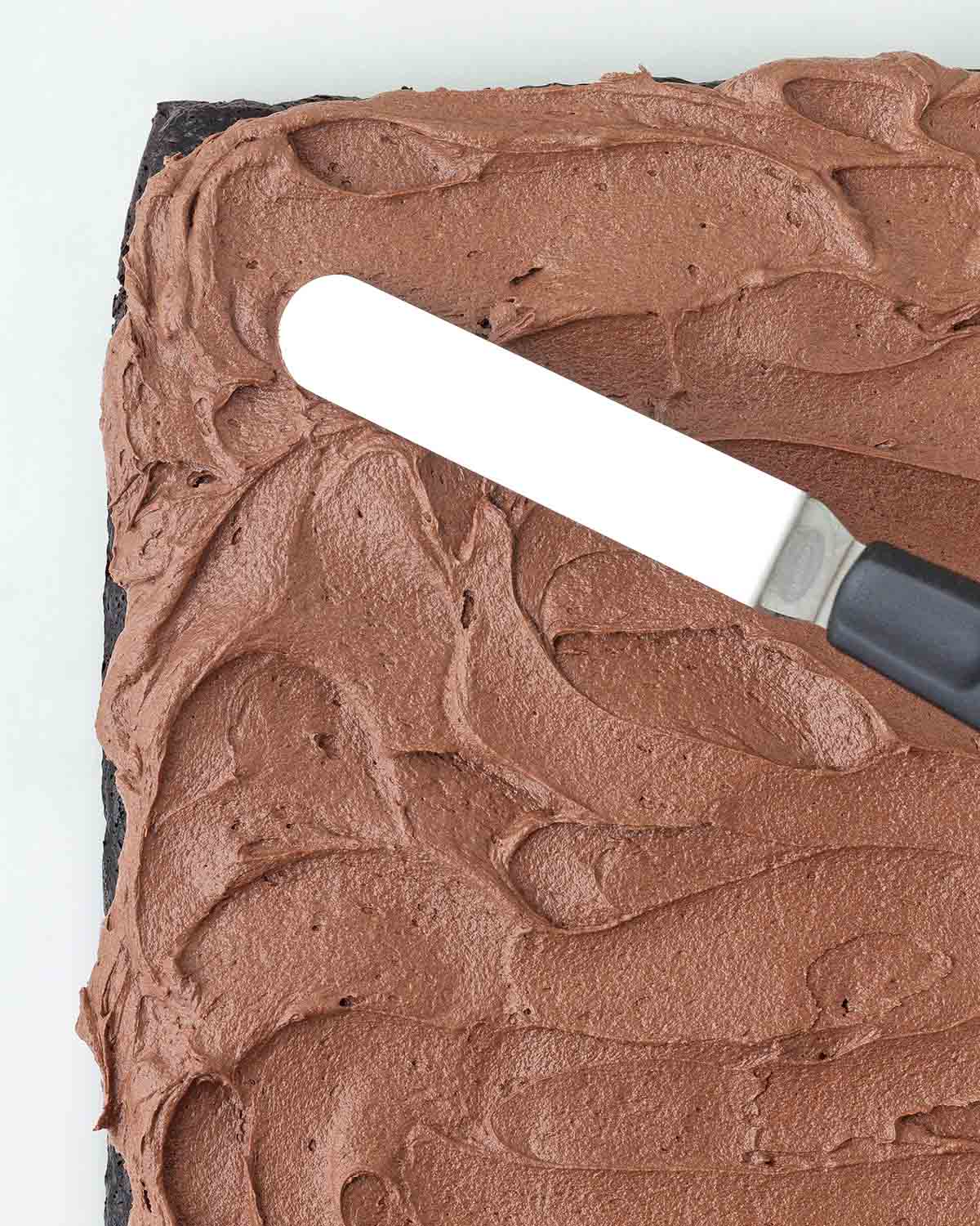 A small offset spatula spreading chocolate frosting on a chocolate sheet cake.