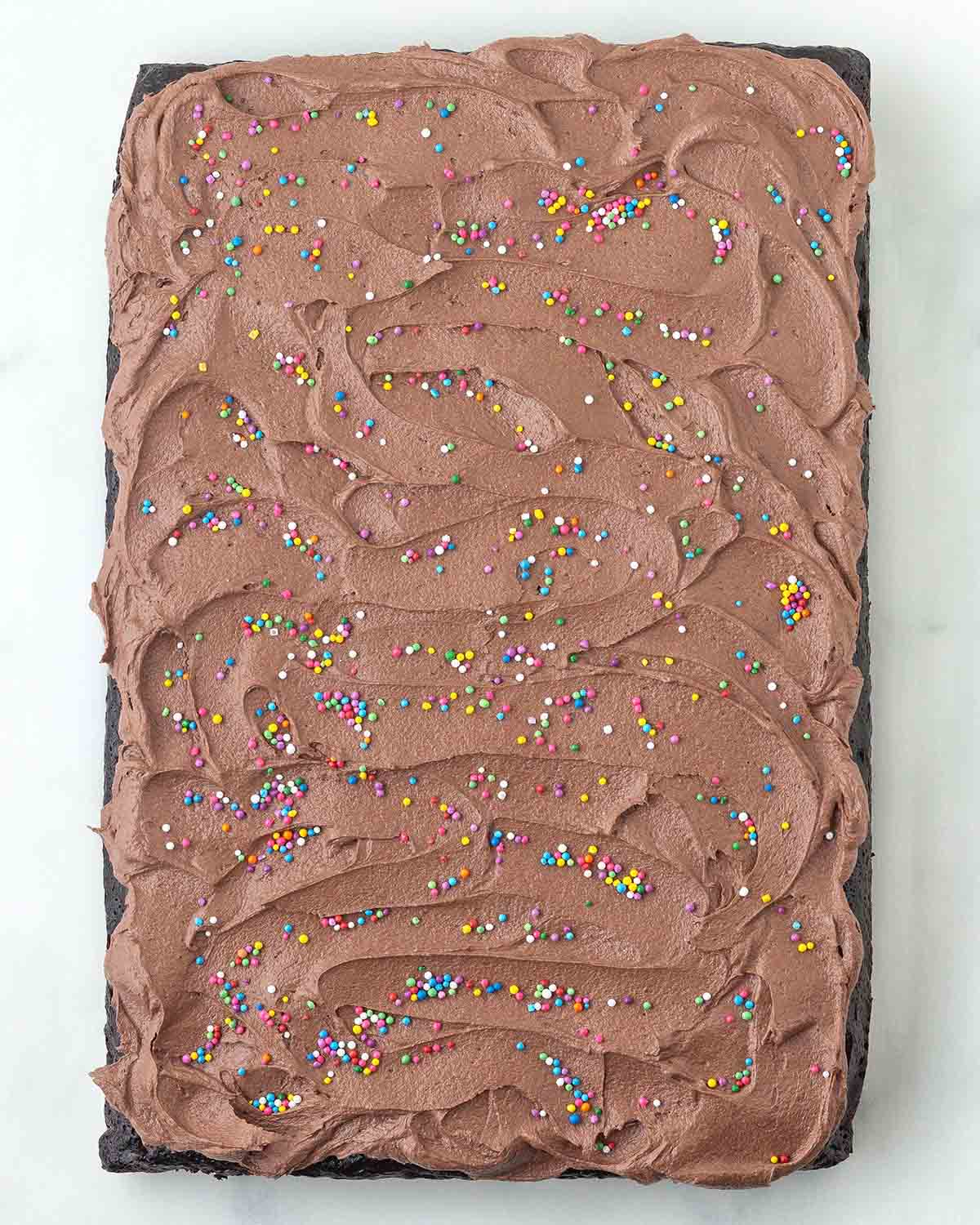 An overhead image showing a chocolate sheet cake that has been topped with chocolate frosting and rainbow sprinkles.