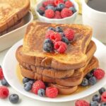 A stack of French toast and berries on a plate, maple syrup has been poured over the stack.