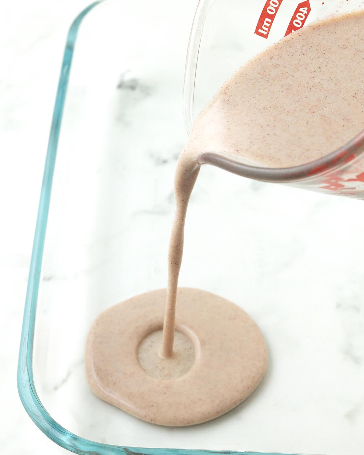 A milk mixture being poured from a measuring cup into a rectangular glass dish.