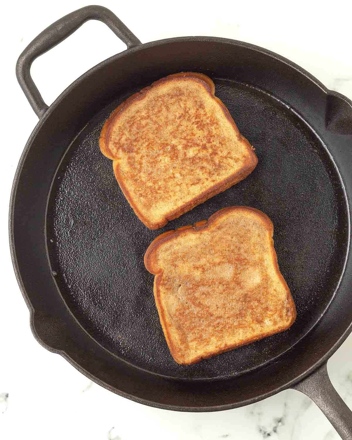 Two pieces of French toast frying in a cast iron skillet.