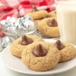 Three peanut butter blossom cookies on a plate, more cookies sit in the back along with a glass of milk.