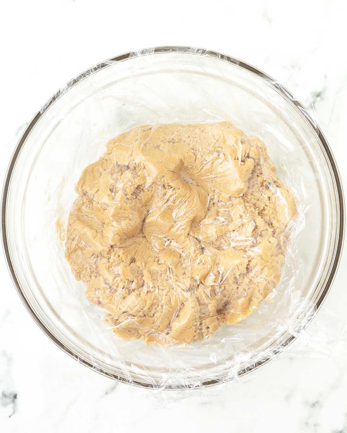 Vegan peanut butter blossom cookie dough in a glass bowl with plastic wrap pressed on top of it just before it is chilled.
