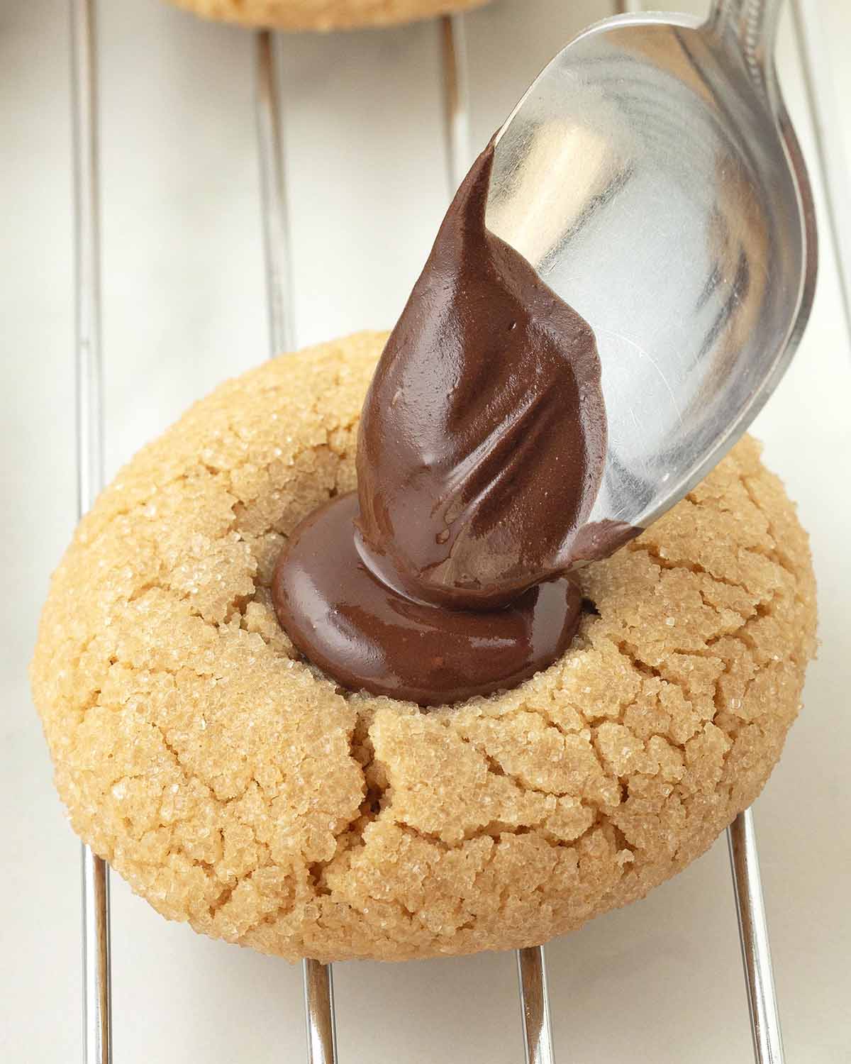 A spoon putting melted chocolate into an indent in a peanut butter cookie.