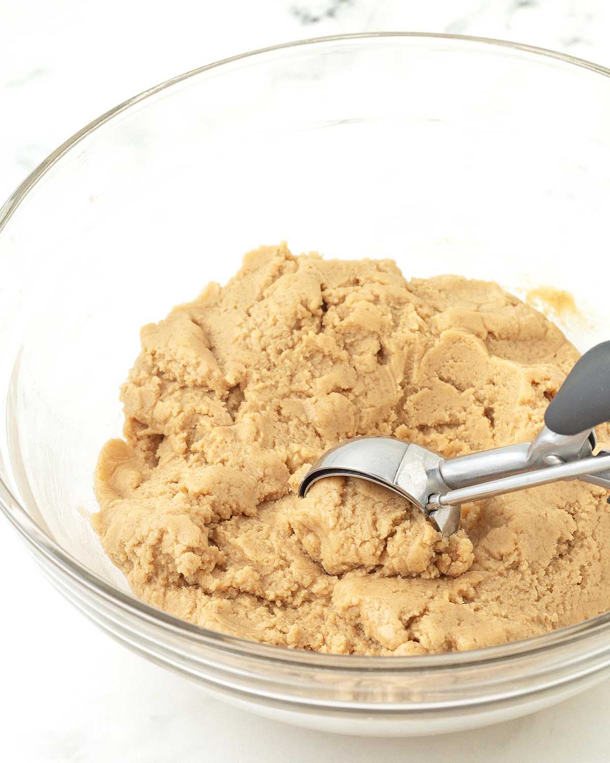 A cookie scoop scooping up cookie dough from inside a glass bowl.