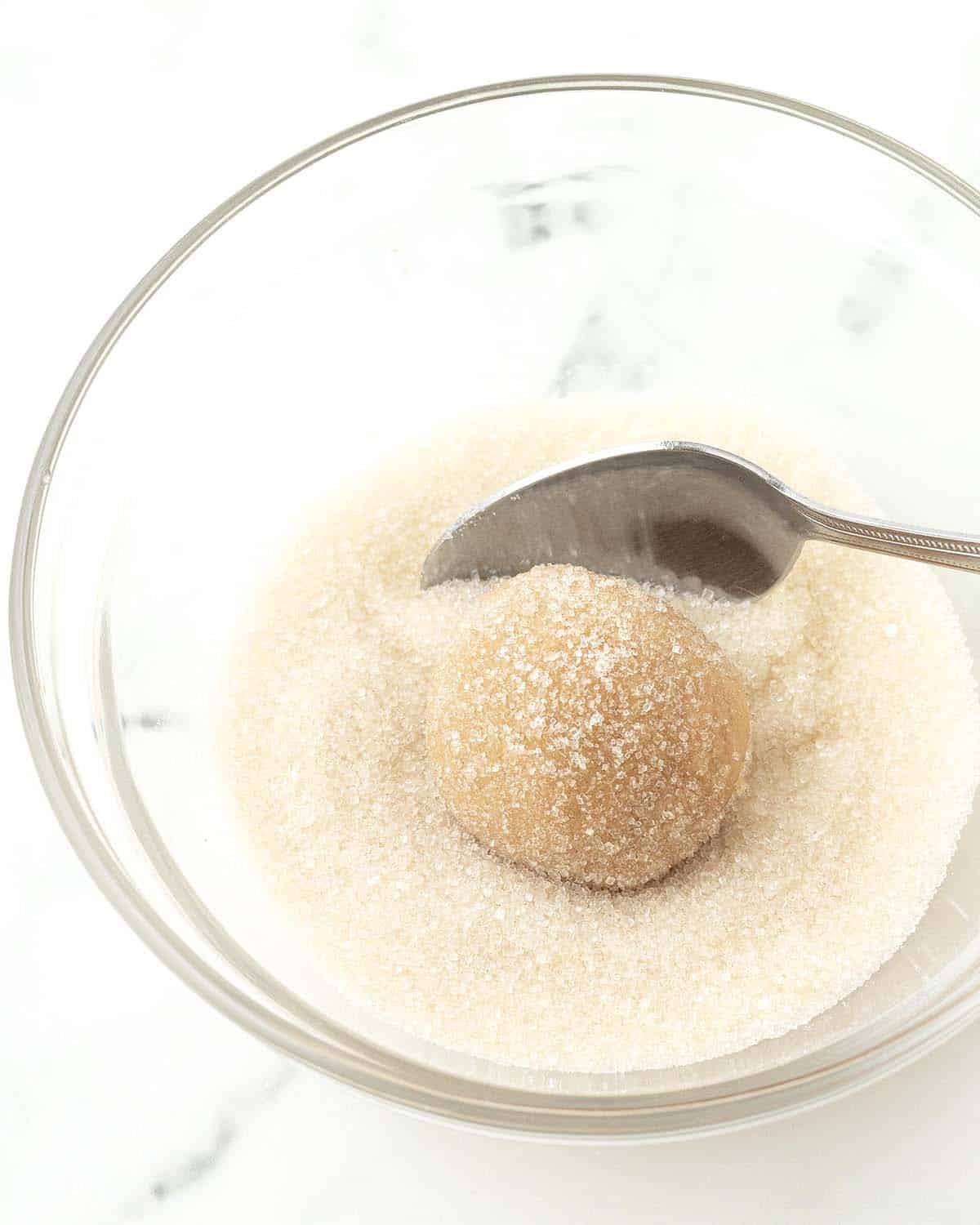 A peanut butter blossom cookie dough ball in a bowl of sugar being rolled around with a spoon to coat it.