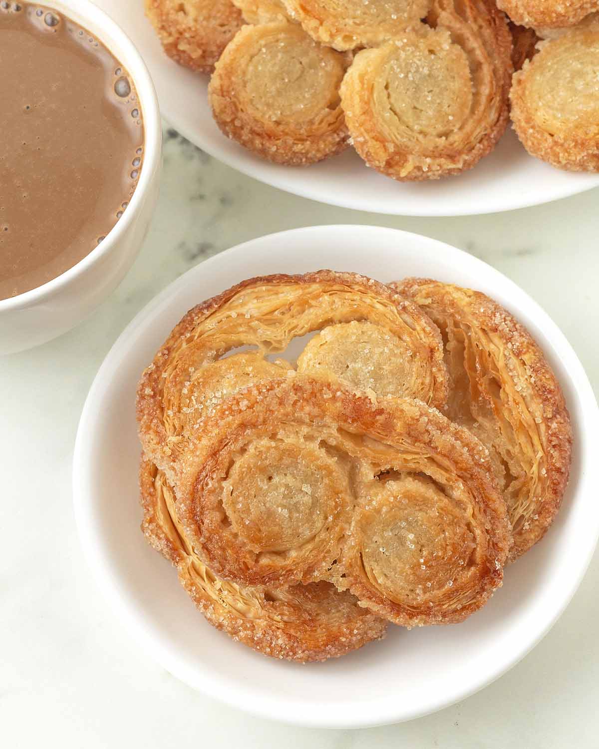 Vegan palmiers on a plate, more palmiers sit on another plate and a mug of hot chocolate sits to the side.