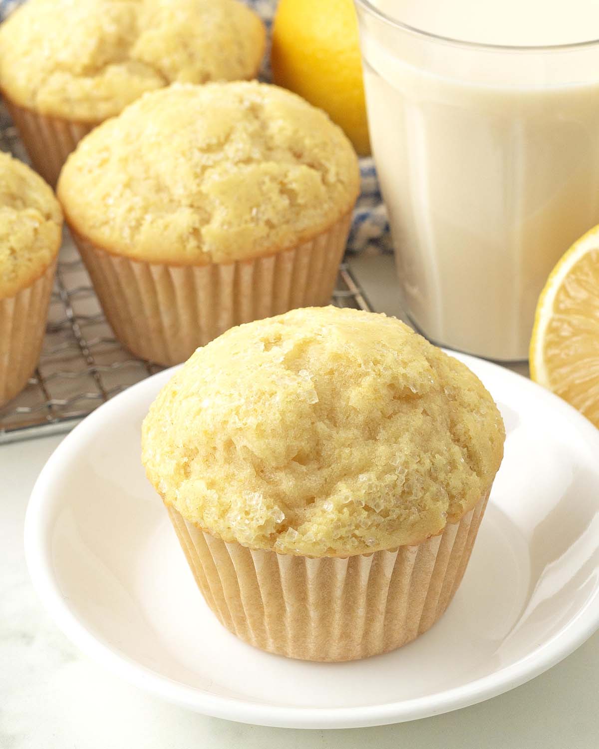 A vegan lemon muffin on a white plate, more muffins, fresh lemons, and a glass of milk are in the background.