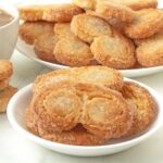 A small plate of vegan puff pastry palmiers sits in the foreground, a larger plate with more pastries sits in the background.