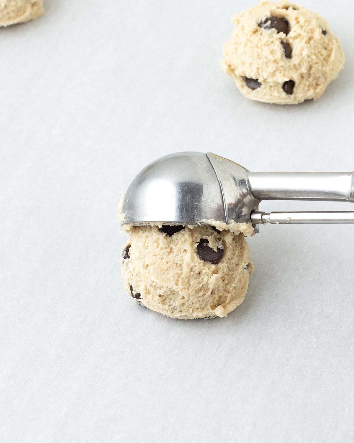 A cookie scoop scooping chocolate chip cookie dough onto a cookie sheet.