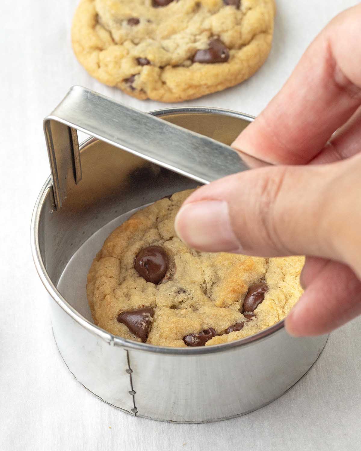 A hand scooting a freshly baked chocolate chip cookie with a biscuit cutter to make it round.