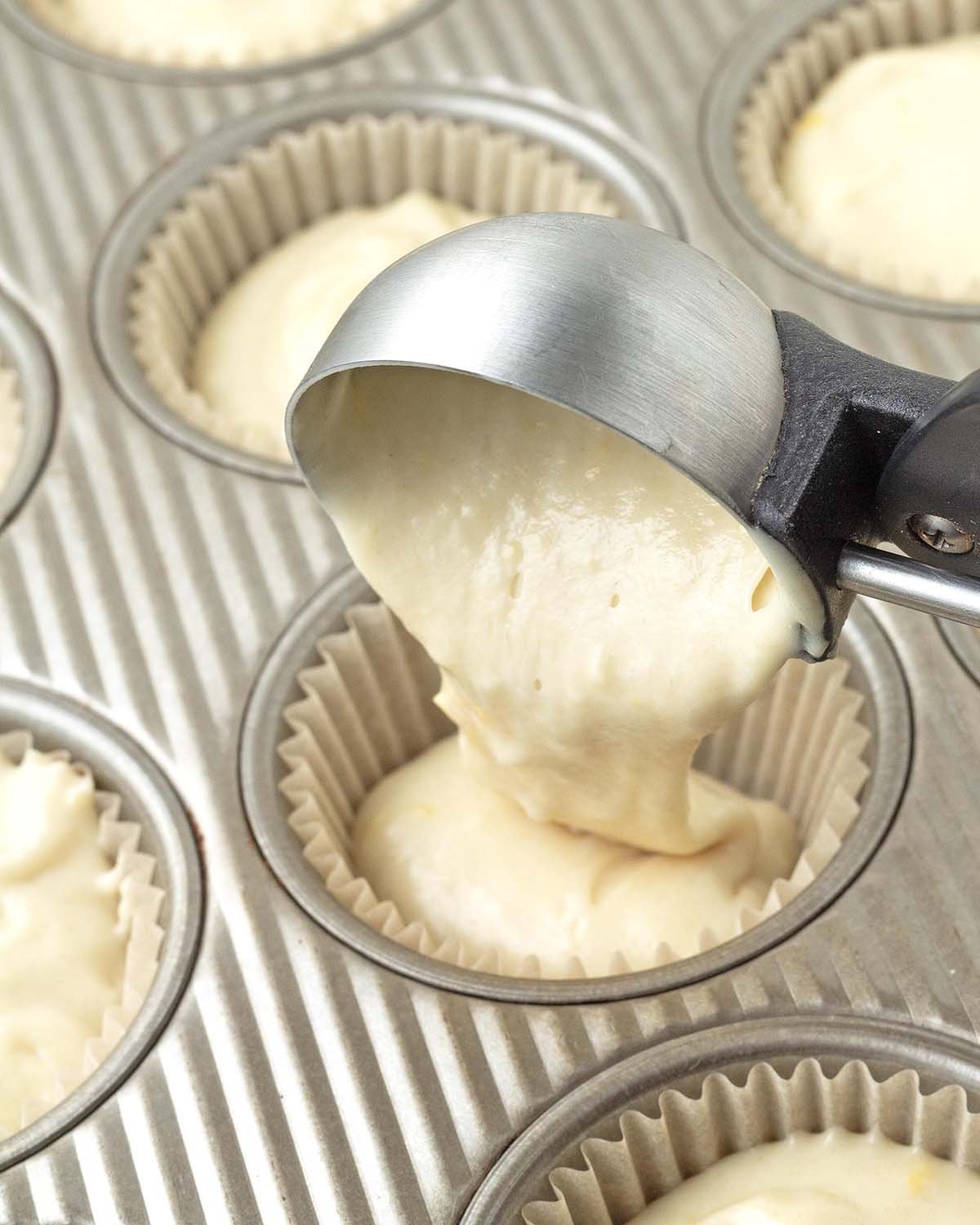 A medium scoop adding lemon muffin batter to a paper muffin cup in a muffin pan.