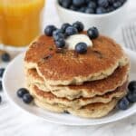 A stack of four blueberry pancakes on a plate, a pat of butter and fresh blueberries sit on top of the stack.