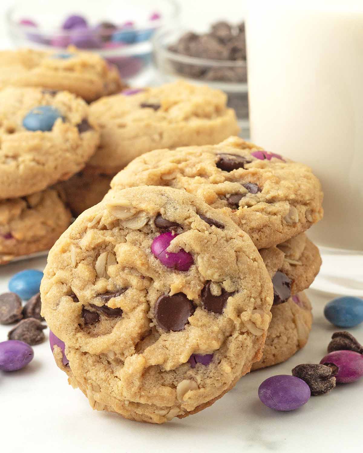 A stack of three monster cookies with one cookie leaning against the stack, more cookies sit in the background.