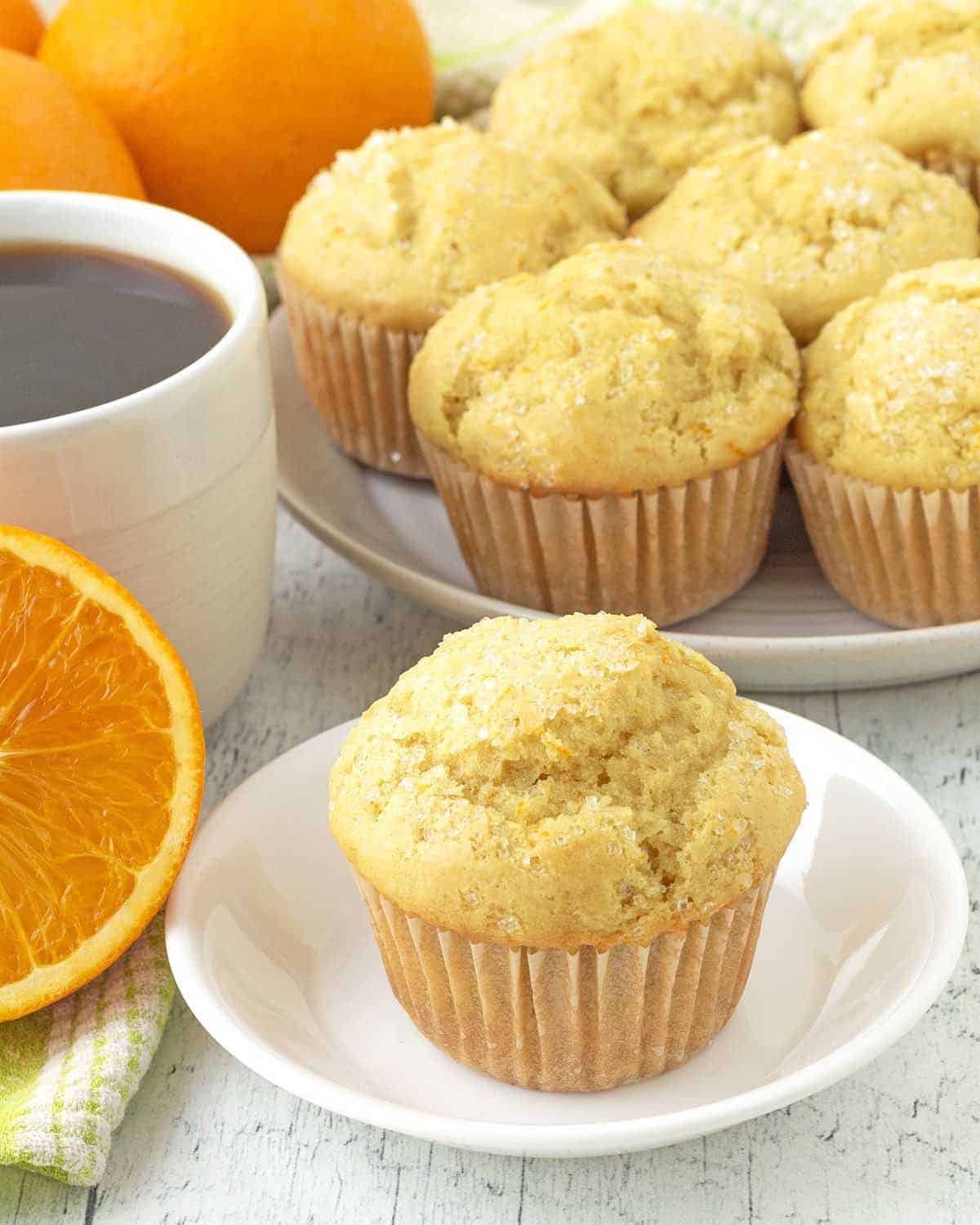 A vegan orange muffin on a white plate, more muffins, fresh oranges, and a cup of coffee are in the background.
