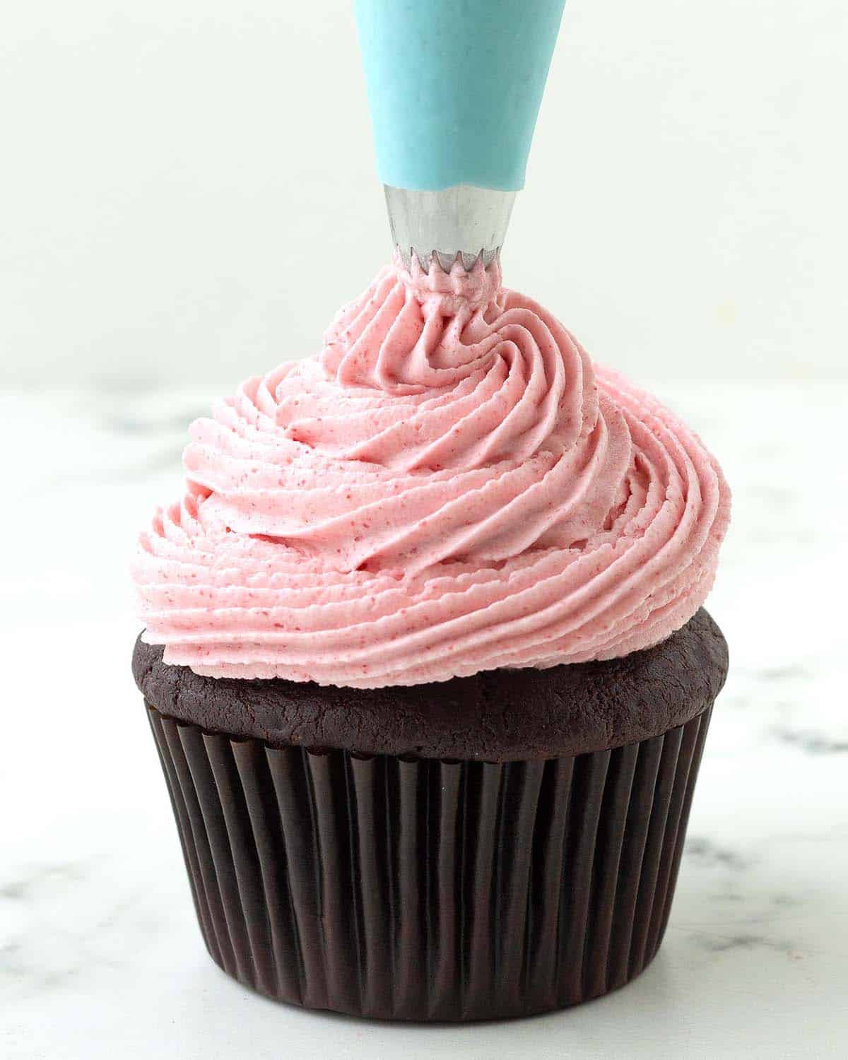 Raspberry frosting being piped onto a chocolate cupcake with a blue coloured piping bag.
