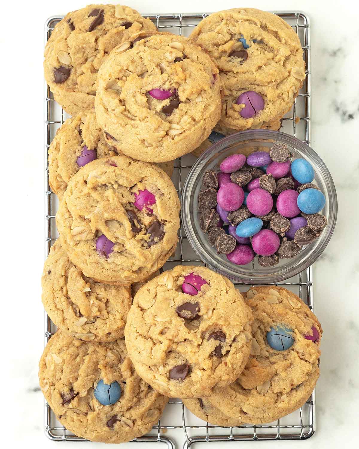 Monster cookies and a small bowl of chocolate candies and chocolate chips on a metal rack.