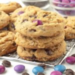 Three vegan monster cookies stacked on top of each other, the stack is sitting on a metal rack with other cookies behind it.