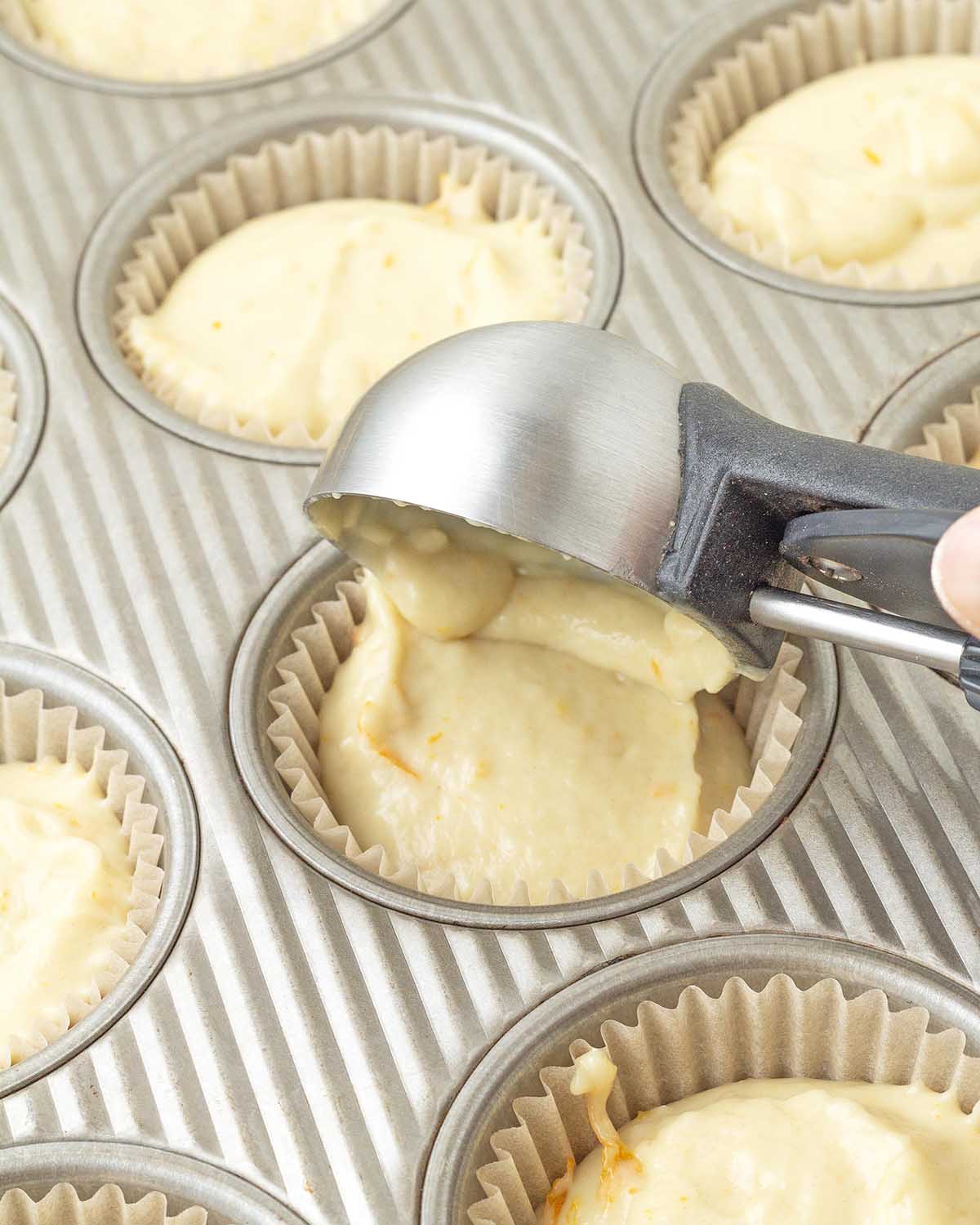 A medium scoop adding orange muffin batter to a paper muffin cup in a muffin pan.