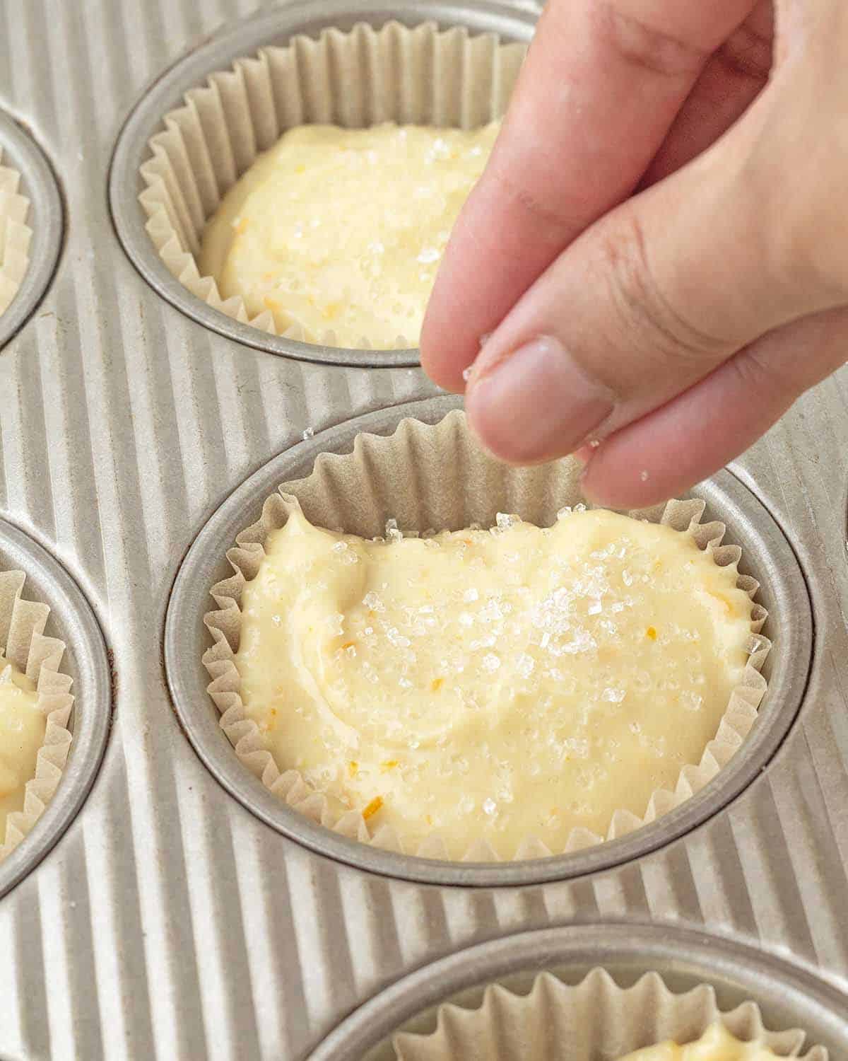 A hand sprinkling coarse sugar on top of an unbaked orange muffin.