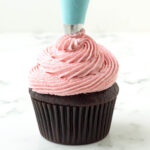 Vegan raspberry buttercream frosting is being piped onto a chocolate cupcake.