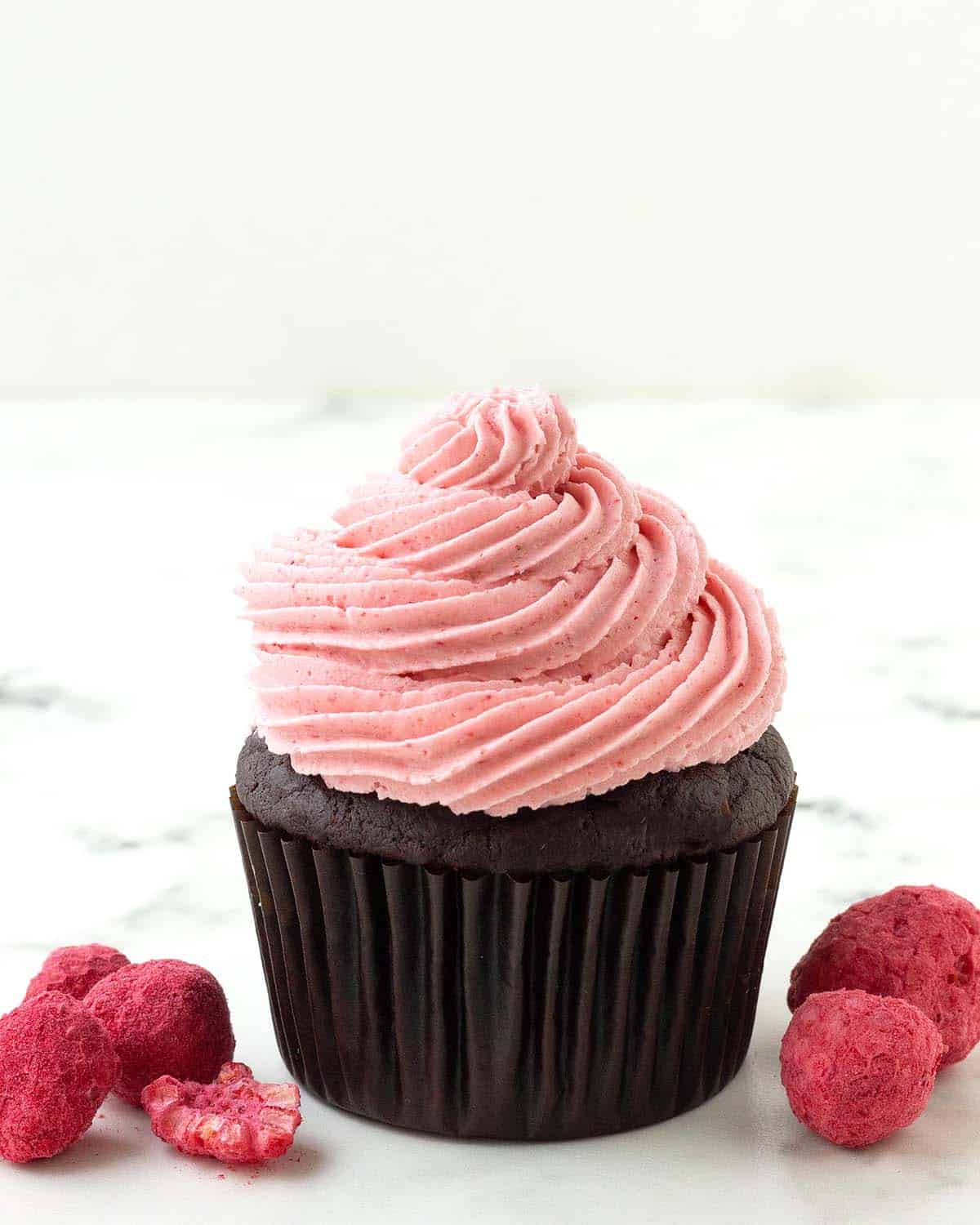 A chocolate cupcake topped with raspberry frosting, freeze dried raspberries sit on both sides of the cupcake.