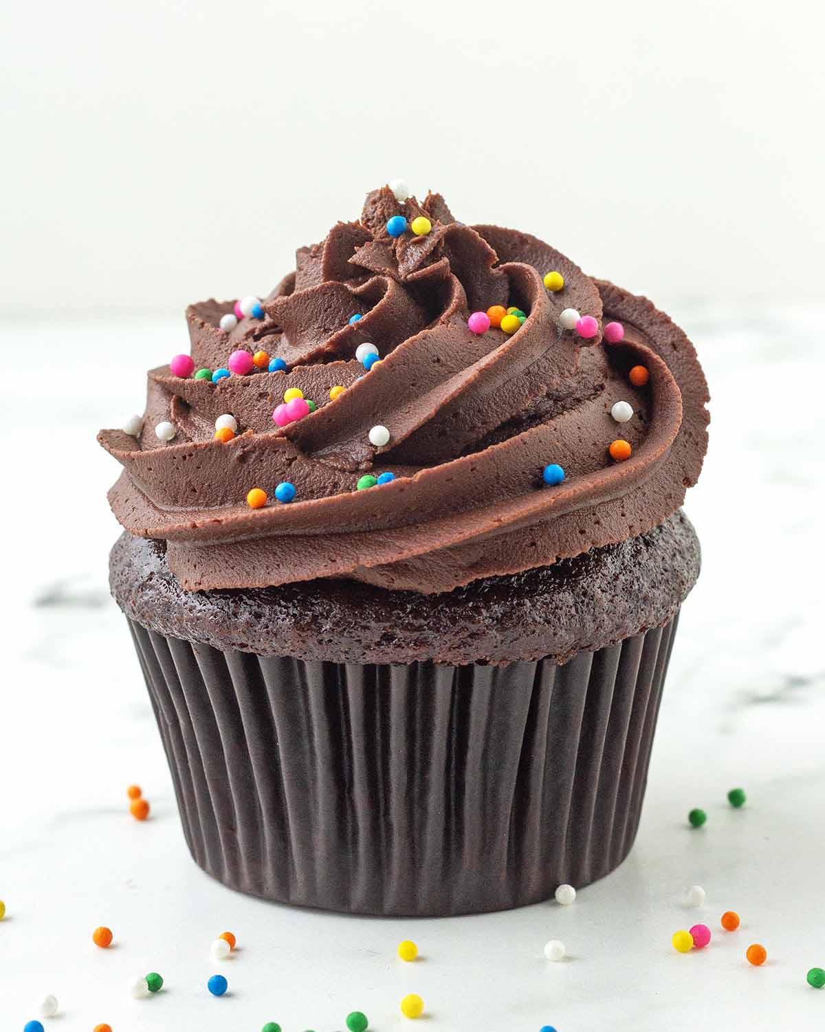 A close up of a frosted chocolate cupcake with round rainbow sprinkles on top and around the cupcake.