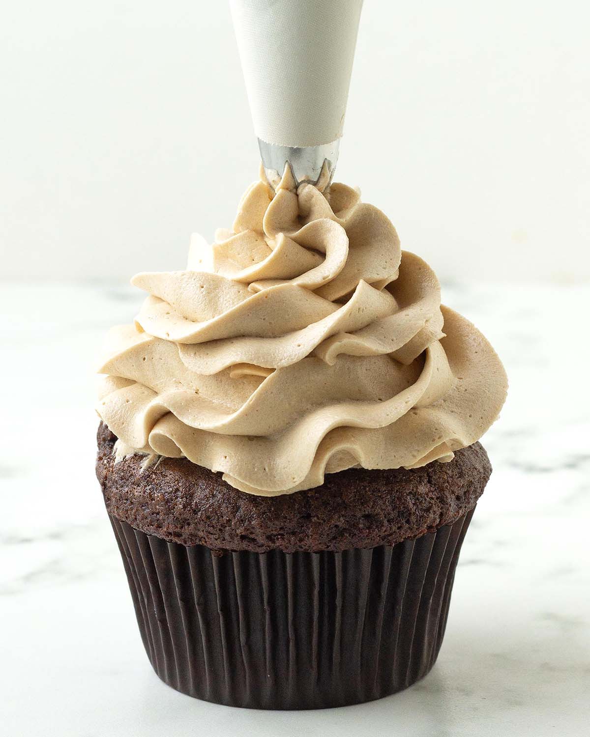 Coffee frosting being piped onto a chocolate cupcake with a white coloured piping bag.