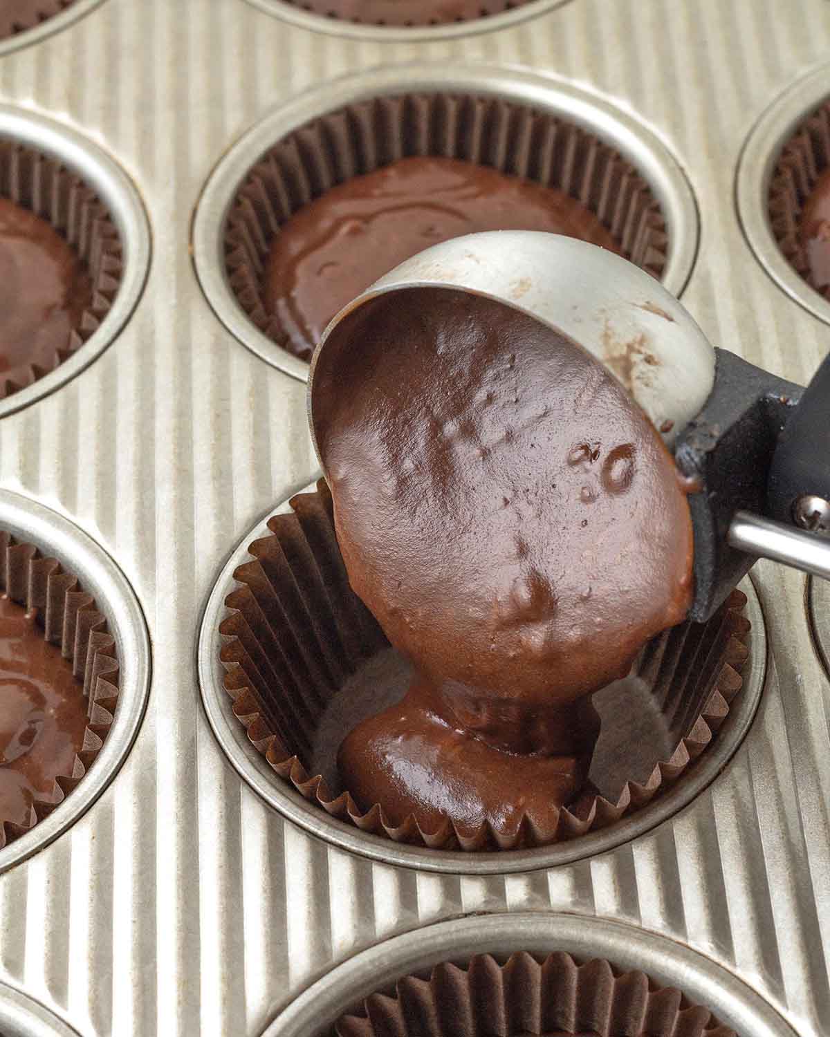 A scoop adding chocolate cupcake batter to a paper cupcake liner that sites in a cupcake pan.