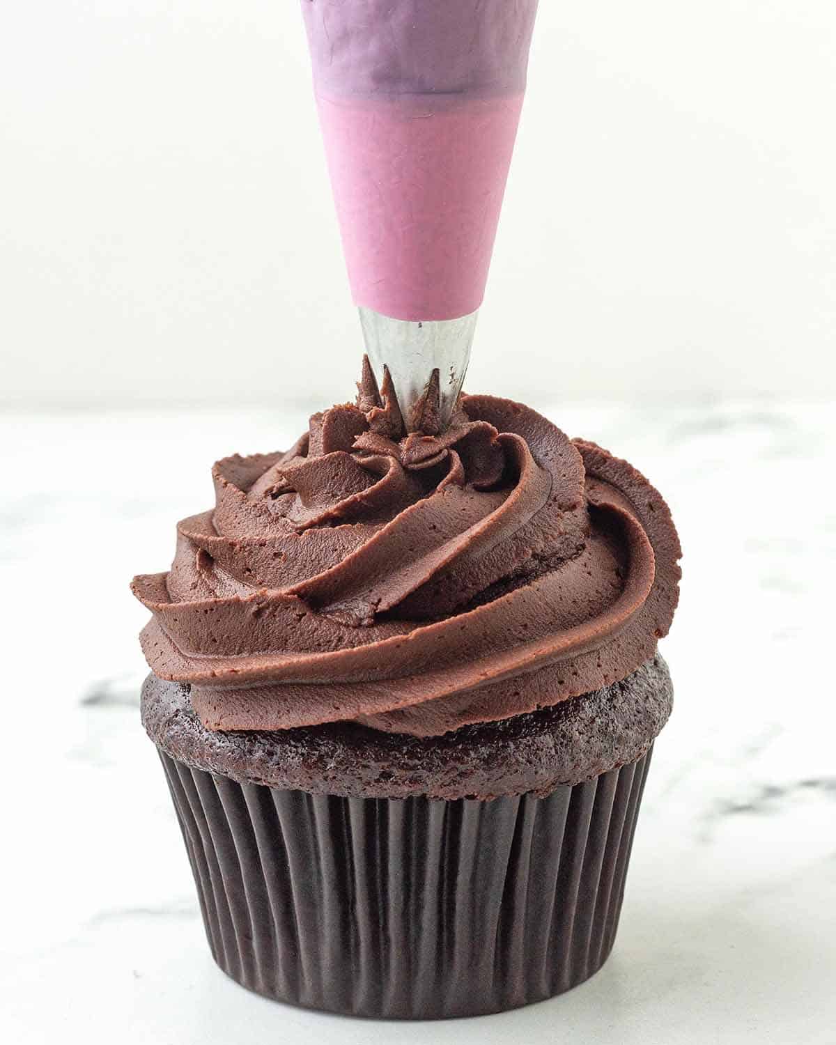 Chocolate ganache frosting being piped onto a chocolate cupcake with a pink coloured piping bag.