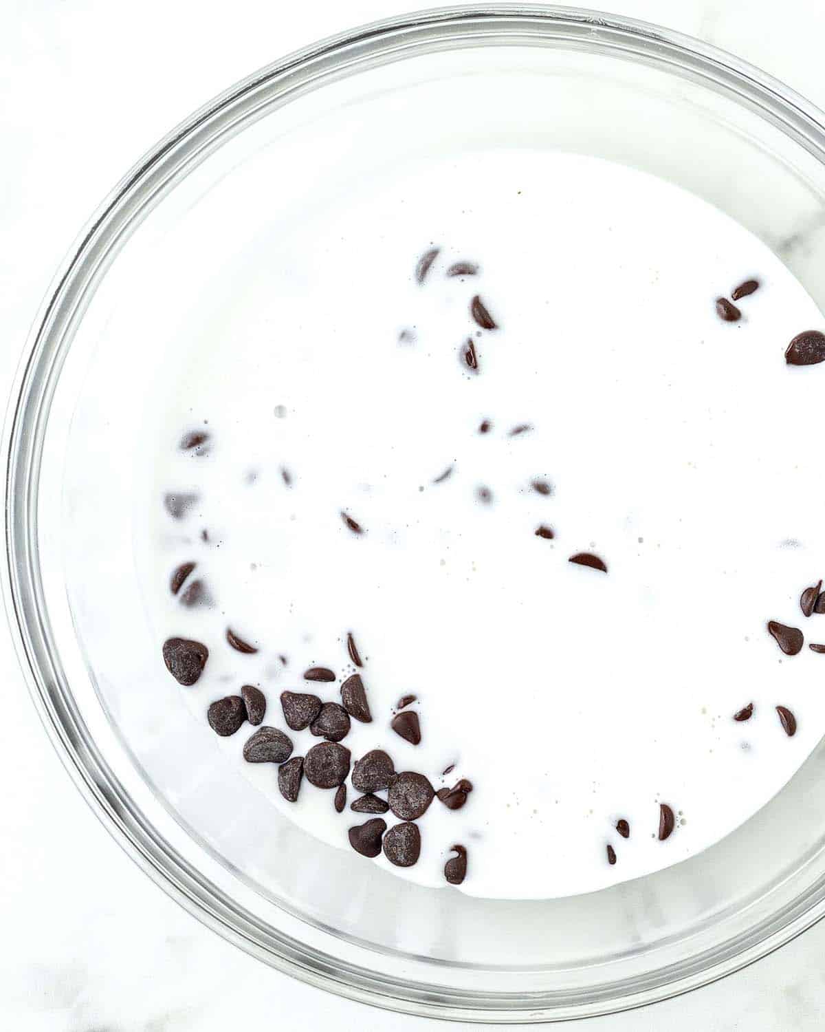 A glass bowl filled with chocolate chips and heavy cream, most of the chocolate chips are under the cream.