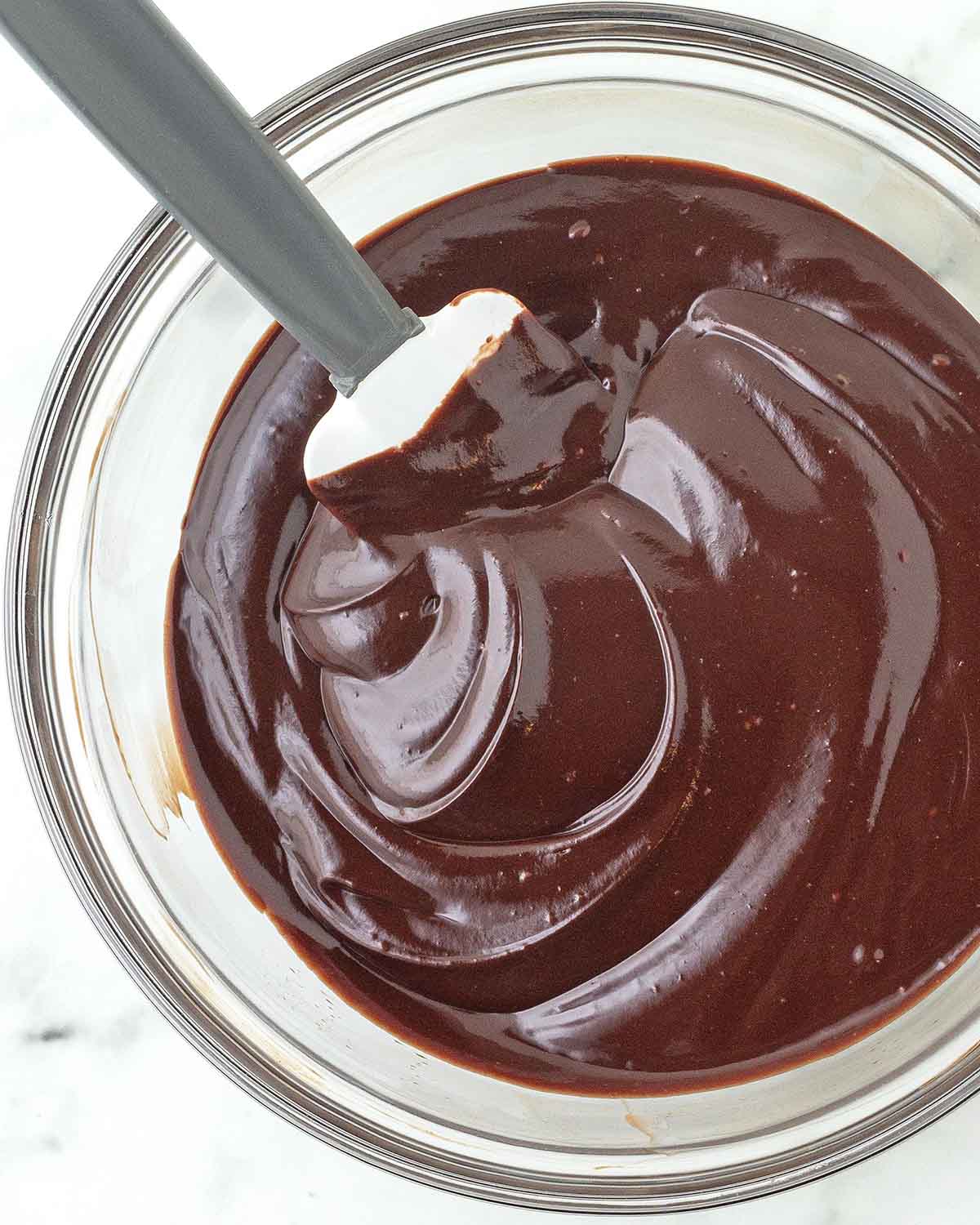 A glass bowl filled with chocolate ganache, the bowl has a rubber spatula in it as well.