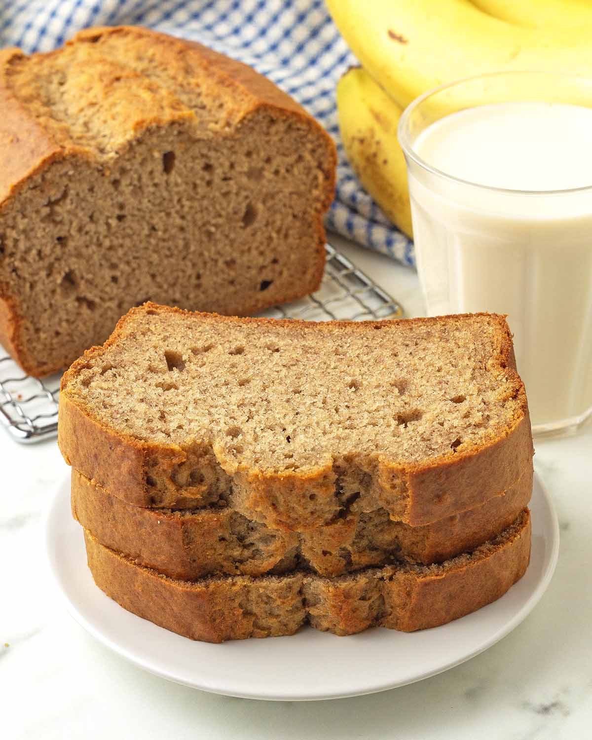 Three slices of banana bread on a plate, the rest of the banana bread, along with a glass of milk and fresh bananas sit in the background.