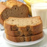 A stack of three slices of banana bread on a plate, the rest of the banana bread, along with a glass of milk sit in the background.