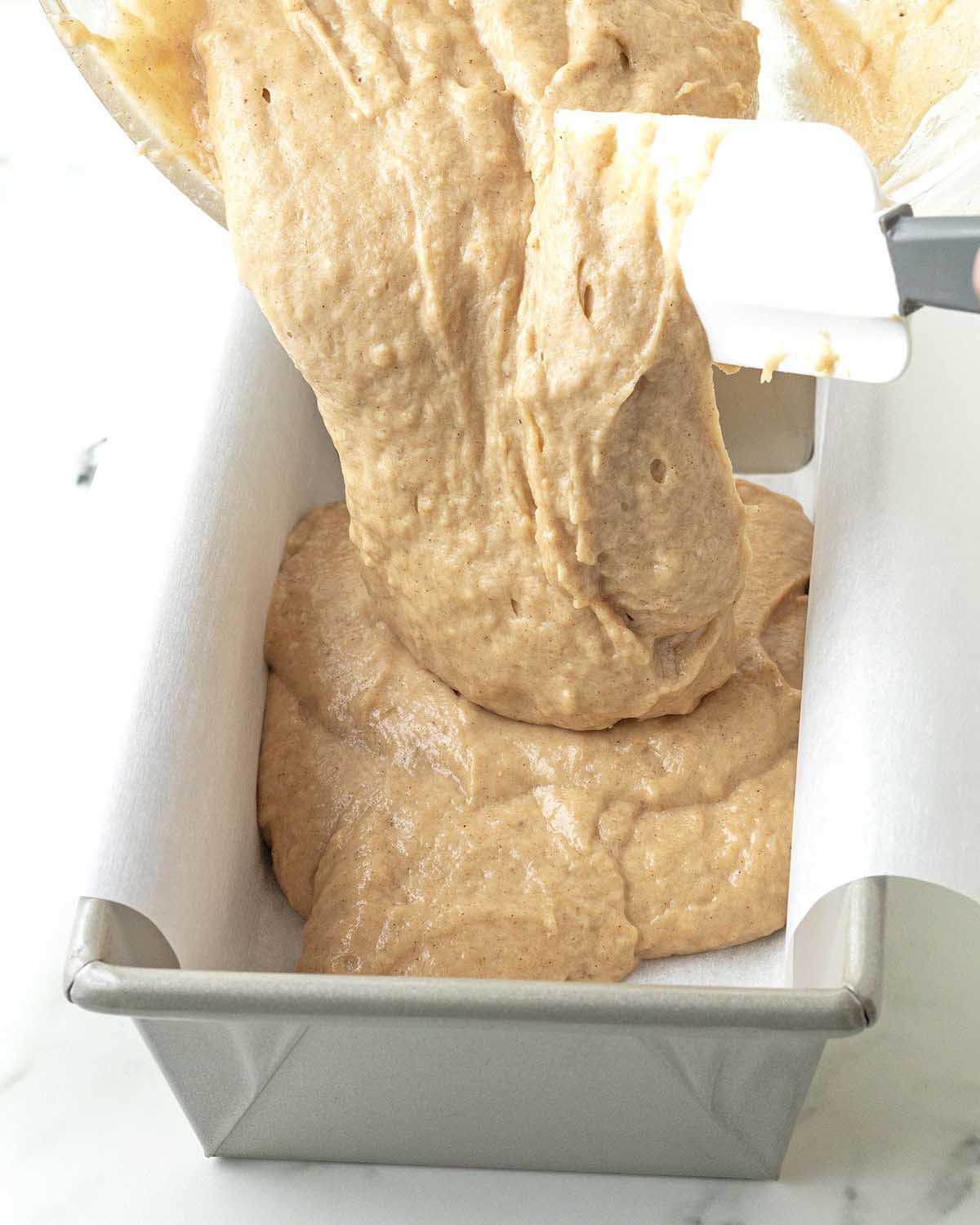 A spatula scraping banana bread batter from a bowl into a loaf pan for baking.