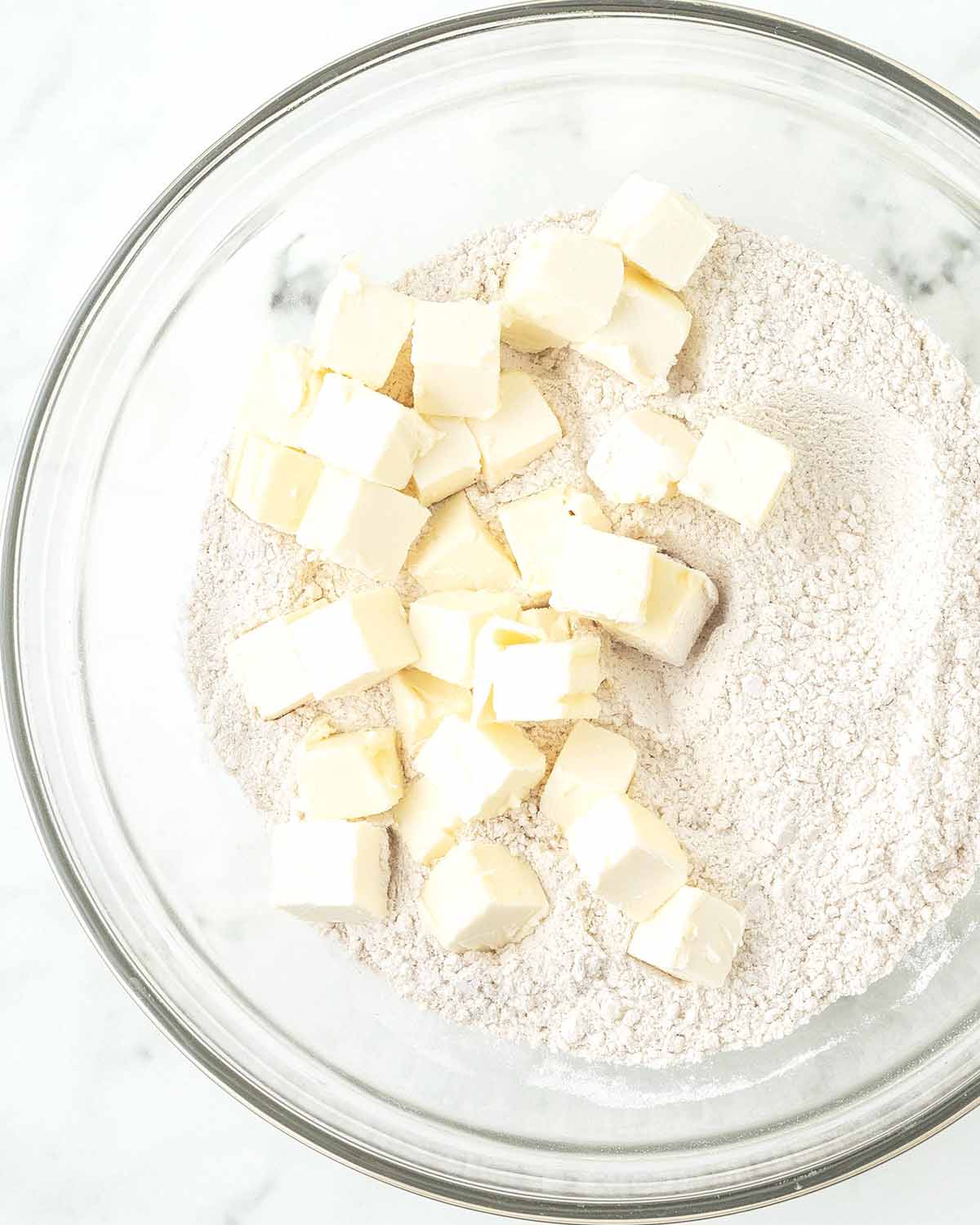 A glass bowl with cubed butter sitting on top of flour.