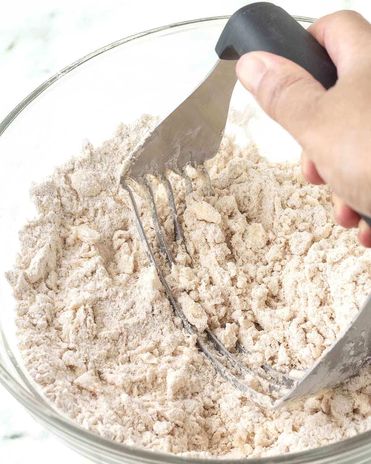 A hand cutting butter into a bowl of dry ingredients using a pastry cutter.