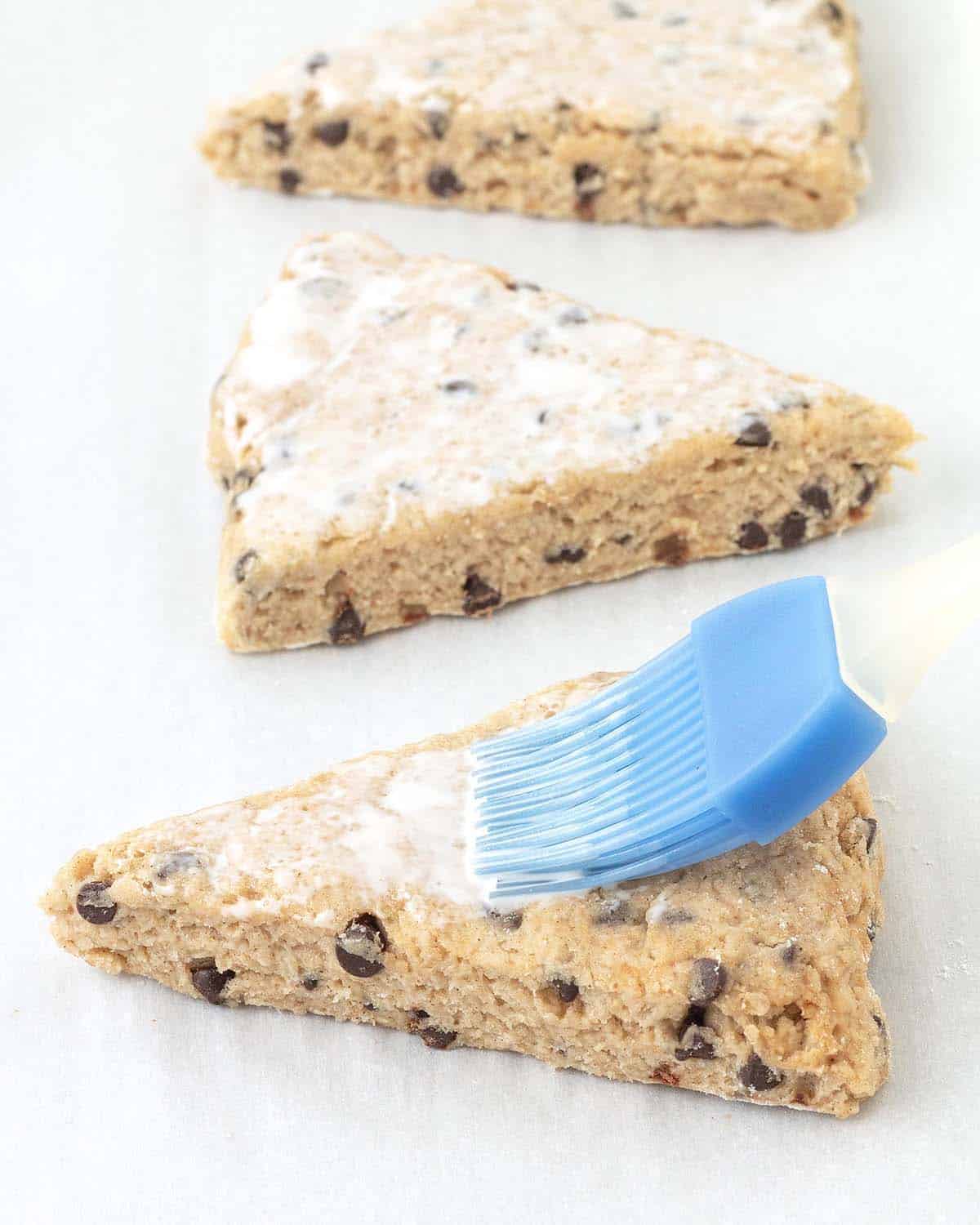 A pastry brush brushing vegan heavy cream on top of an unbaked chocolate chip scone.