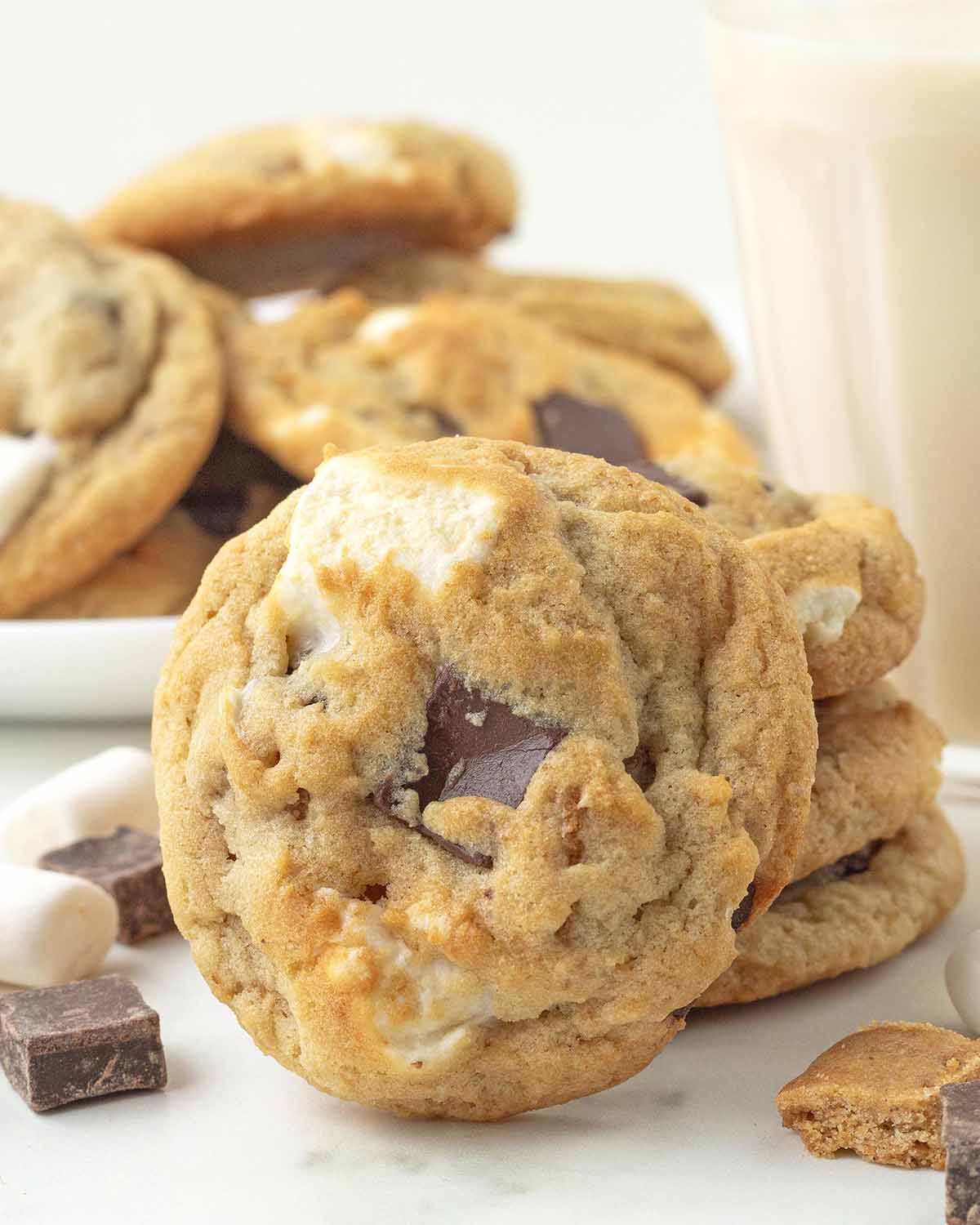 A stack of three s'mores cookies with one cookie leaning against the stack, more cookies sit in the background.