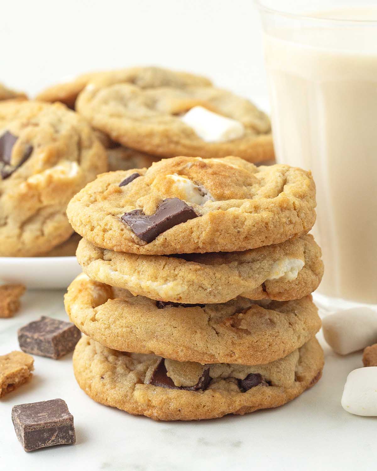 A stack of four s'mores cookies with chocolate, marshmallows, and graham crackers around it, more cookies sit in the background.