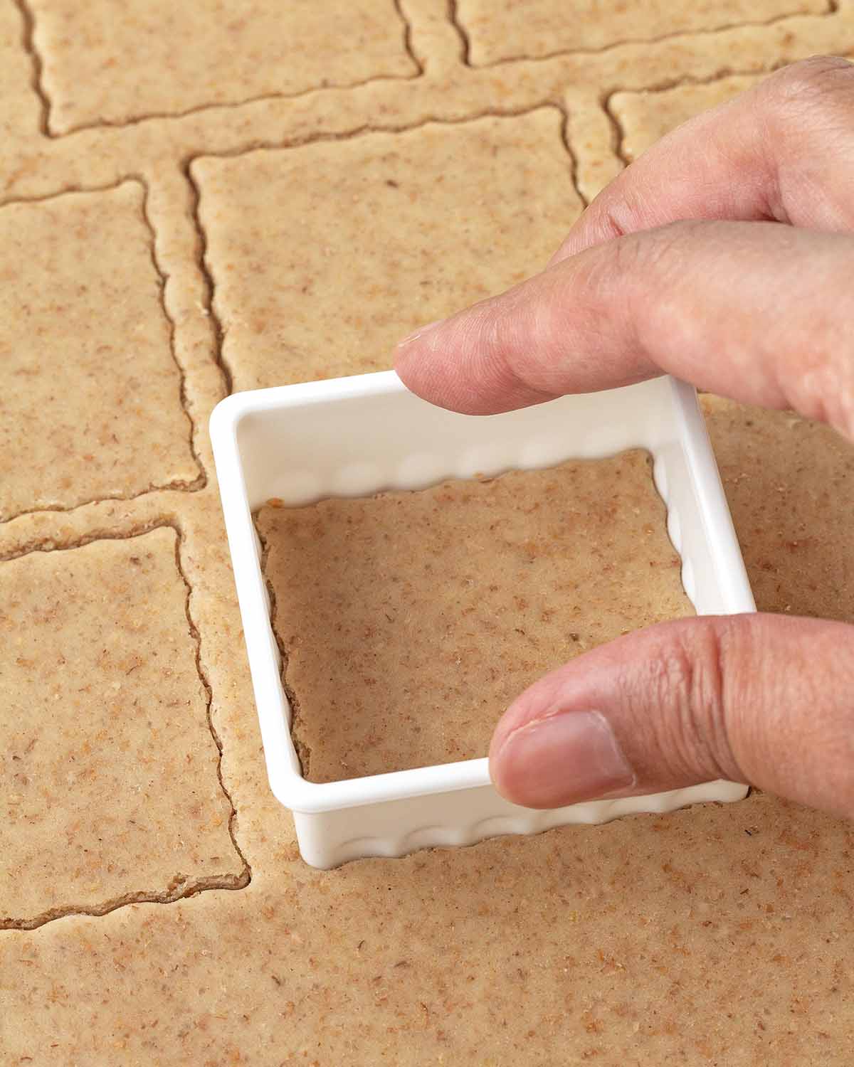 A hand pressing a square cookie cutter into rolled graham cracker dough.