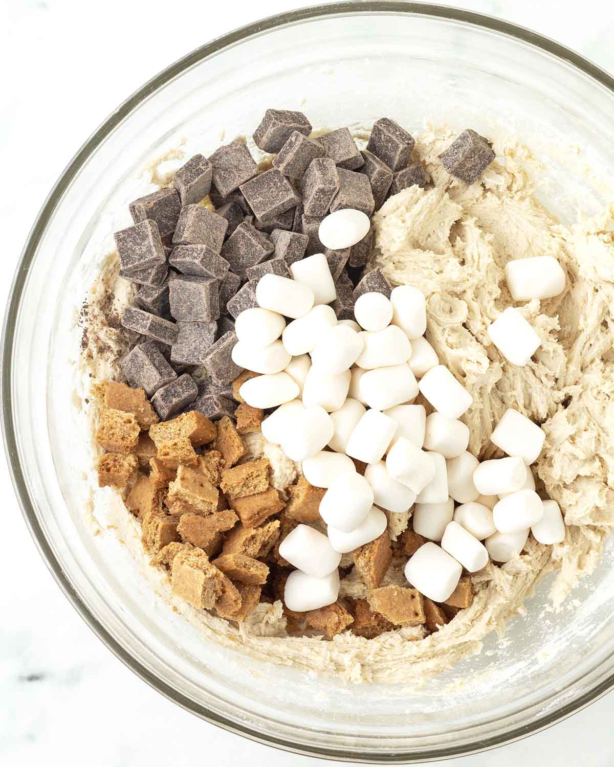 Smores cookie dough in a bowl with chocolate, marshmallows, and graham cracker pieces on top waiting to be mixed in.