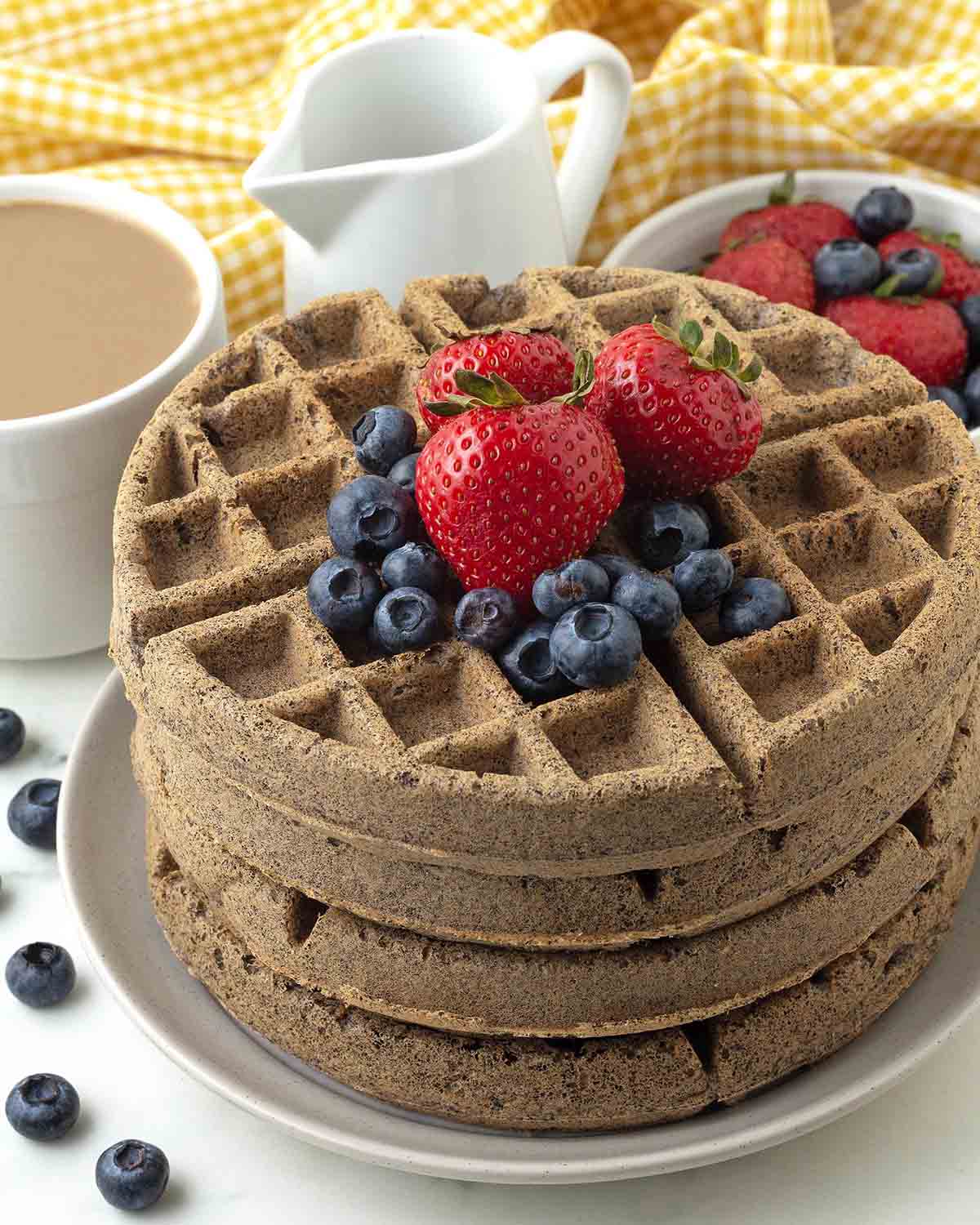 Four buckwheat waffles stacked on top of each other on a white plate, the stack is topped with fresh berries.