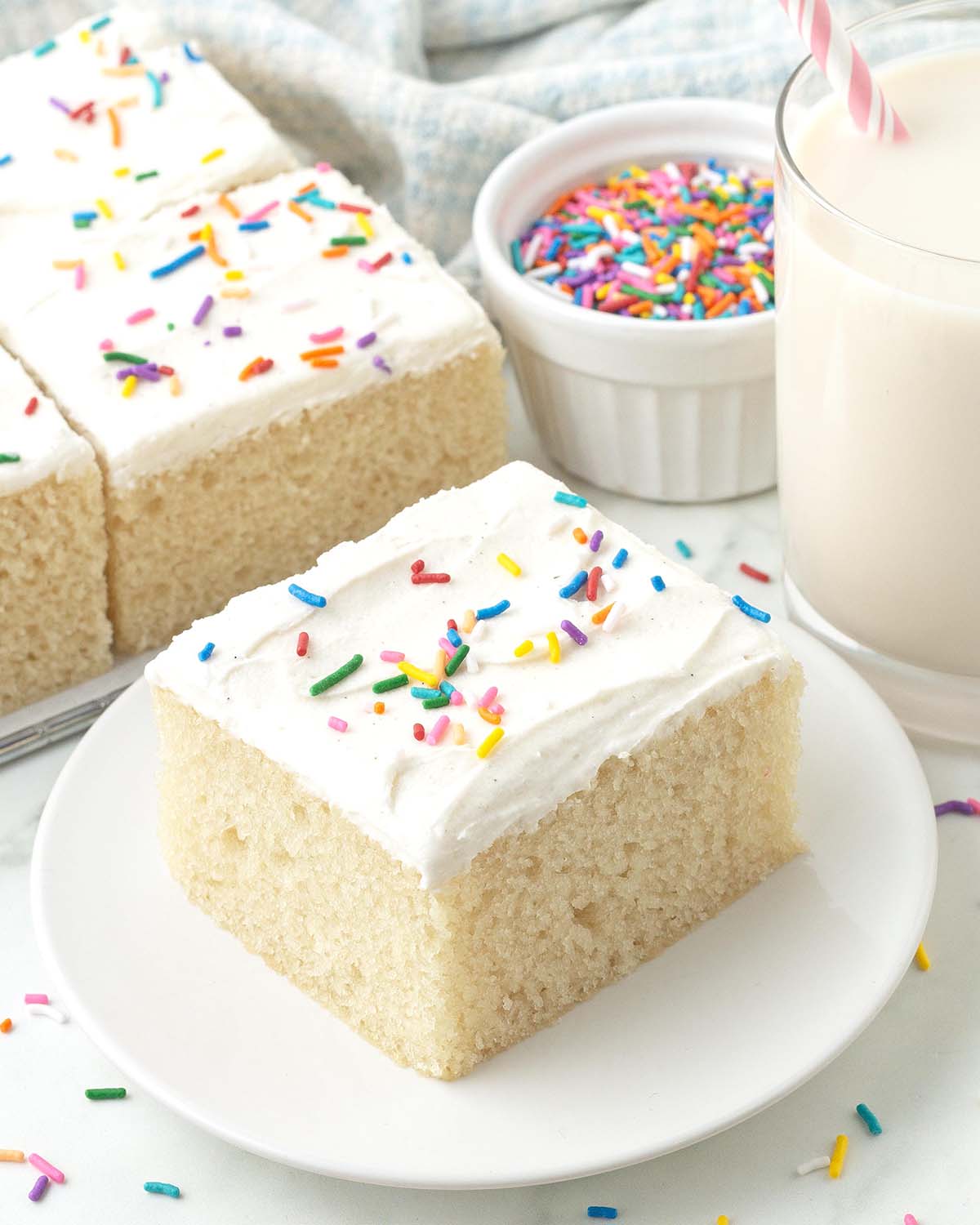 A square of frosted vegan vanilla sheet cake with sprinkles sitting on a plate.