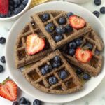 An overhead image of a buckwheat waffle on a plate, fresh berries and syrup top the waffle.